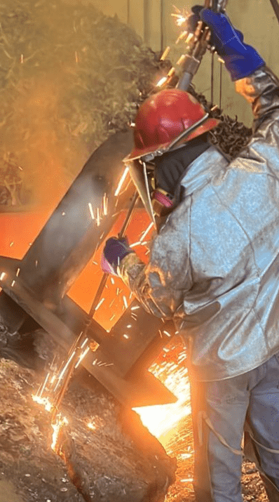 IMX worker wearing protective gear cutting a large metal object with a torch held overhead.